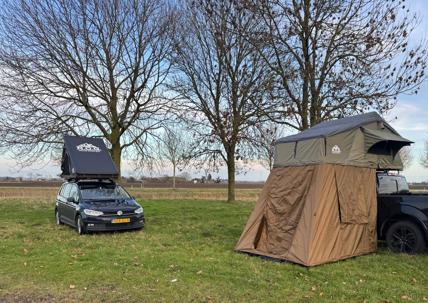 Kampeeropstelling met tent op pickup, twee auto’s op grasveld, kale bomen en bewolkte lucht.